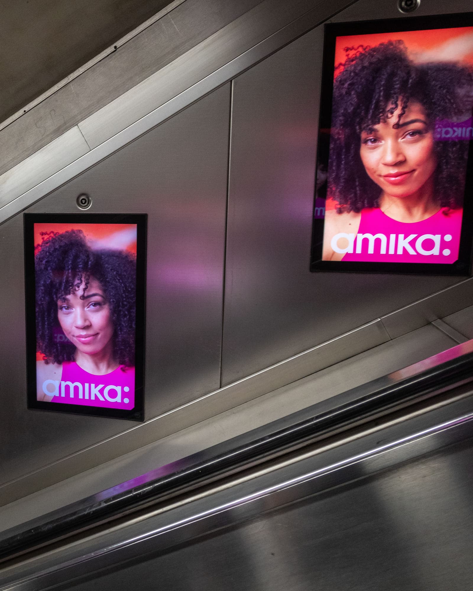 Two digital escalator panels carrying the same amika advertisement on the wall beside an Underground escalator.
